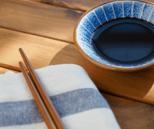 Un plato pequeño de cerámica con salsa de soja junto a unos palillos de madera sobre una mesa de madera.