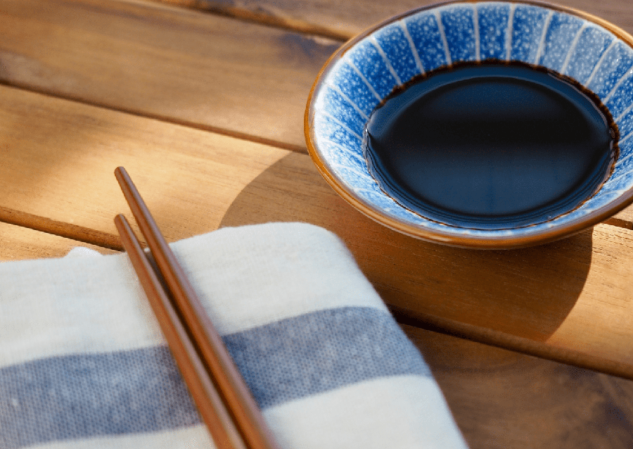 Un plato pequeño de cerámica con salsa de soja junto a unos palillos de madera sobre una mesa de madera.